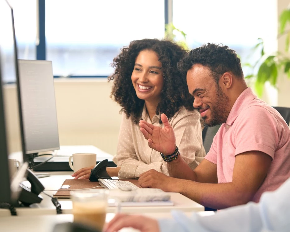 Happy man with down syndrome smiling and sitting at desk with support worker while both looking at computer